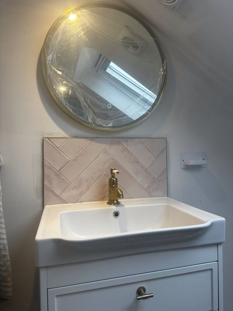 Blush pink herringbone tile splashback behind a white vanity unit with a brass tap